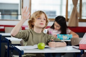 Schoolboy Looking Away While Raising Hand At Desk